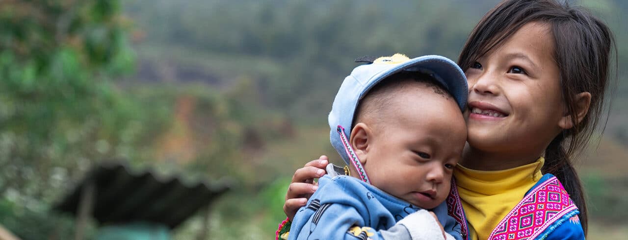Zwei Kinder lachen und kuscheln im Freien. Hintergrund ist eine verschwommene, natürliche Landschaft.