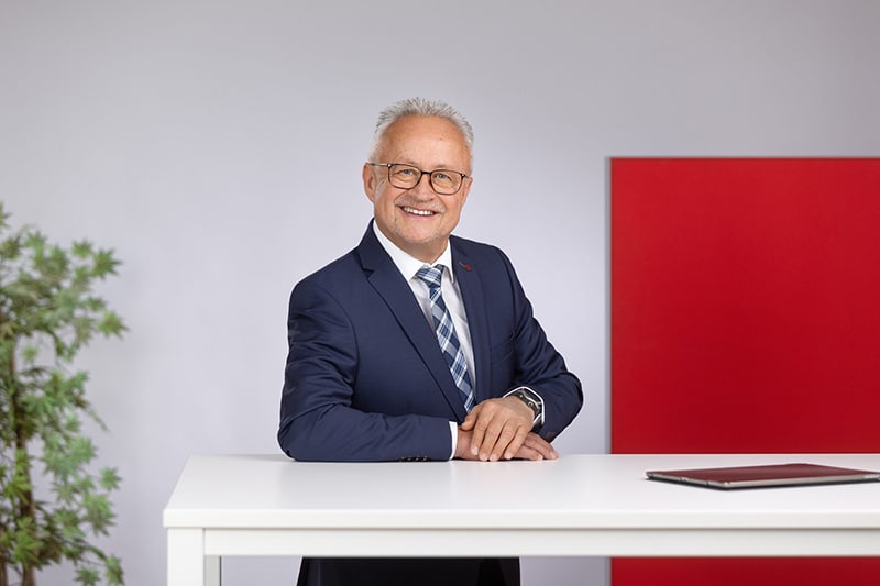 Mitarbeiter in Anzug steht am Tisch einer Bürofirma mit rotem Hintergrund und Pflanzen.