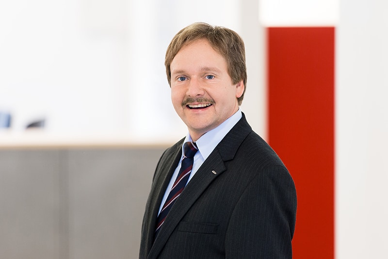 Ein Mitarbeiter in Anzug lächelt in die Kamera. Moderne Büroumgebung von Schröter GmbH im Hintergrund.