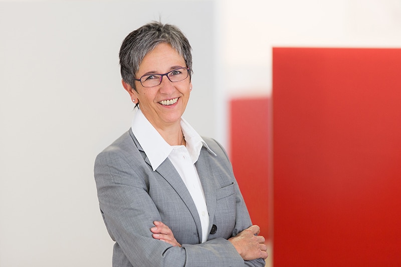 Personalleiterin der Schröter GmbH in grauem Anzug lächelt vor rotem Hintergrund. C-Artikel-Management.