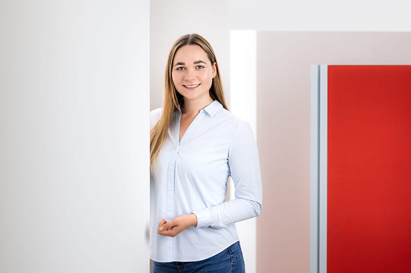Mitarbeiterin in weißem Hemd lächelt freundlich in die Kamera, stehend vor modernem Hintergrund bei Schröter GmbH.