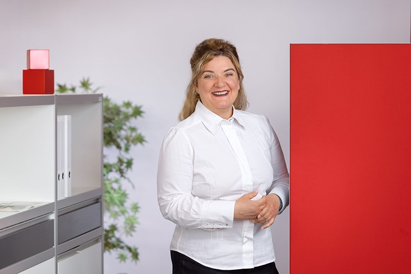 Mitarbeiterin vor rotem Hintergrund, Büroeinrichtung am Rand. Schröter GmbH, C-Artikel-Beschaffung.