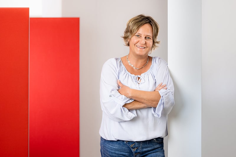 Frau lehnt lächelnd an Wand, roter Hintergrund, symbolisiert Schröter GmbH's C-Artikel-Beschaffungslösungen.
