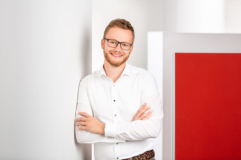 Mitarbeiter im weißen Hemd lächelt vor rotem Hintergrund, repräsentiert Schröter GmbH, Experte für C-Artikel-Beschaffung.