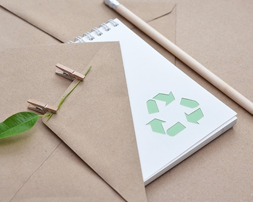 Notizbuch mit Recycling-Symbol, umgeben von Papier, Stift und grünem Blatt; Symbol für nachhaltige Büropraktiken.