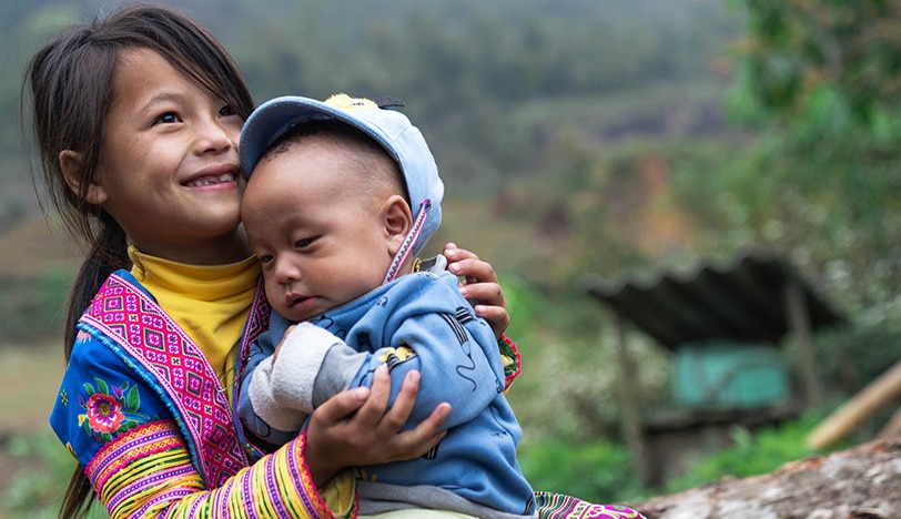 Lächelndes Kind hält Baby in traditioneller Kleidung im Freien. Schöter engagiert sich für Nachhaltigkeit u. a. bei Plan International.