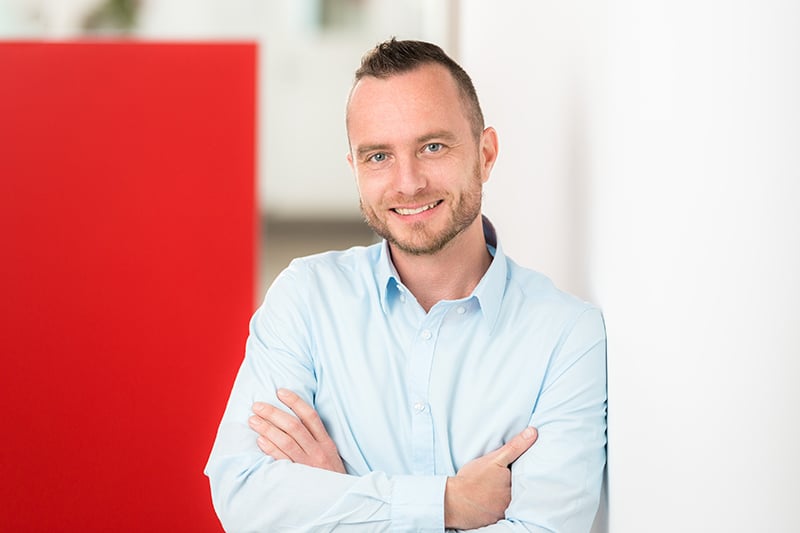 Mitarbeiter lächelt, steht vor rotem Hintergrund; effizienter C-Artikel-Management bei Schröter GmbH.