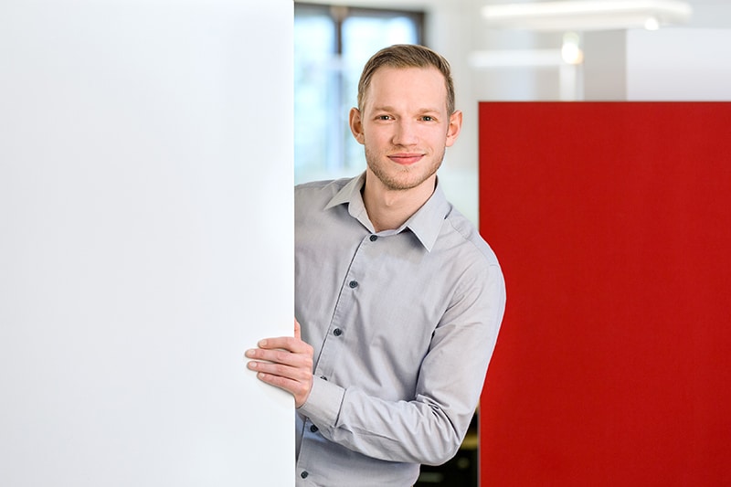 Mitarbeiter von Schröter GmbH lächelt in einem modernen Büro, hinter einer Wand stehend.
