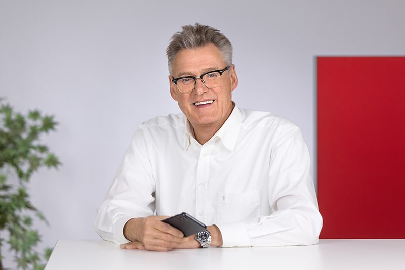 Mitarbeiter im weißen Hemd, hält ein Smartphone, sitzt an einem Tisch. Hintergrund mit rot und grün. Schröter GmbH Thema.